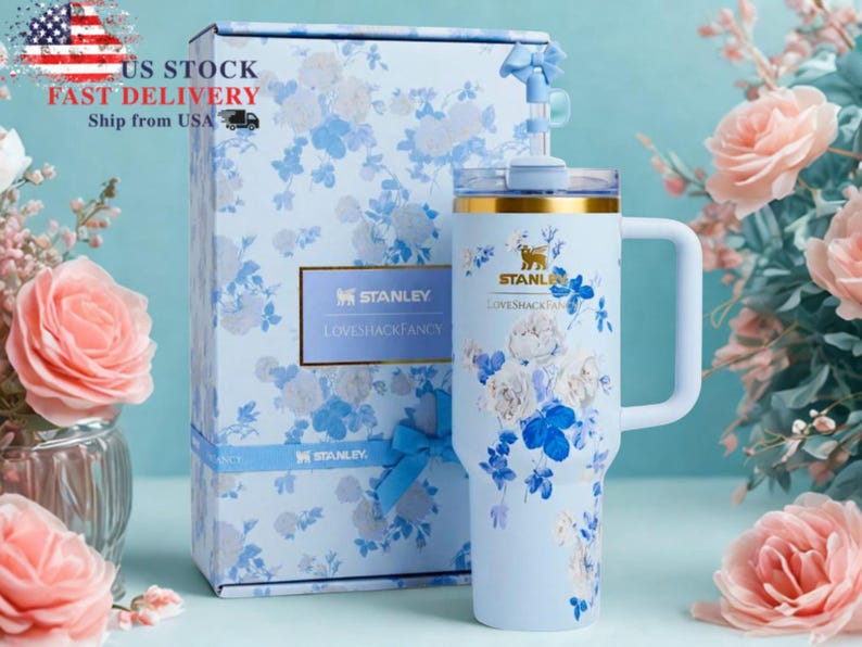 May include: A light blue Stanley tumbler and matching gift box, both adorned with a floral pattern in white and blue. The tumbler has a handle and a gold rim. The box has a blue ribbon and the text "US STOCK FAST DELIVERY Ship from USA".