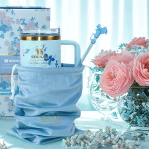 May include: A light blue Stanley mug with a handle, gold trim, and floral design, nestled in a matching drawstring bag. A matching box with a floral pattern is behind the mug. A blue straw and a bouquet of pink roses complete the scene.