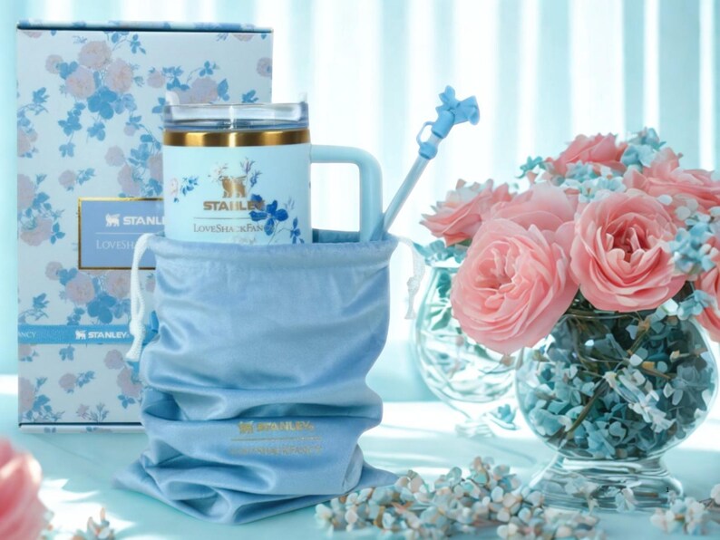 May include: A light blue Stanley mug with a handle, gold trim, and floral design, nestled in a matching drawstring bag. A matching box with a floral pattern is behind the mug. A blue straw and a bouquet of pink roses complete the scene.