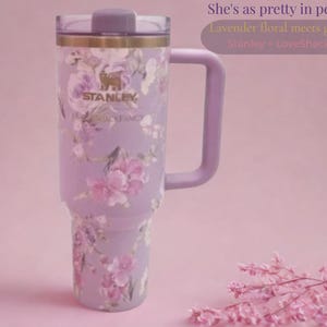 May include: A lavender Stanley tumbler with a floral pattern, a gold-toned rim, and a matching handle. The cup features the text Stanley and LoveShackFancy. The text in the image reads: She's as pretty in person, Lavender floral meets gold detail, Stanley > LoveShackFancy.