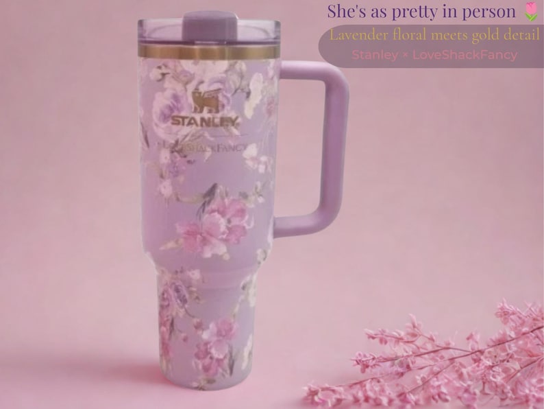 May include: A lavender Stanley tumbler with a floral pattern, a gold-toned rim, and a matching handle. The cup features the text Stanley and LoveShackFancy. The text in the image reads: She's as pretty in person, Lavender floral meets gold detail, Stanley > LoveShackFancy.