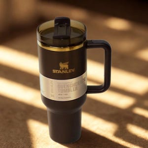 Może przedstawiać: Czarny kubek Stanley Quencher H2.0 z uchwytem i przezroczystą pokrywką. Kubek ma złoty pasek i logo Stanley. Tekst "QUENCHER TUMBLER" jest wydrukowany na białej etykiecie.