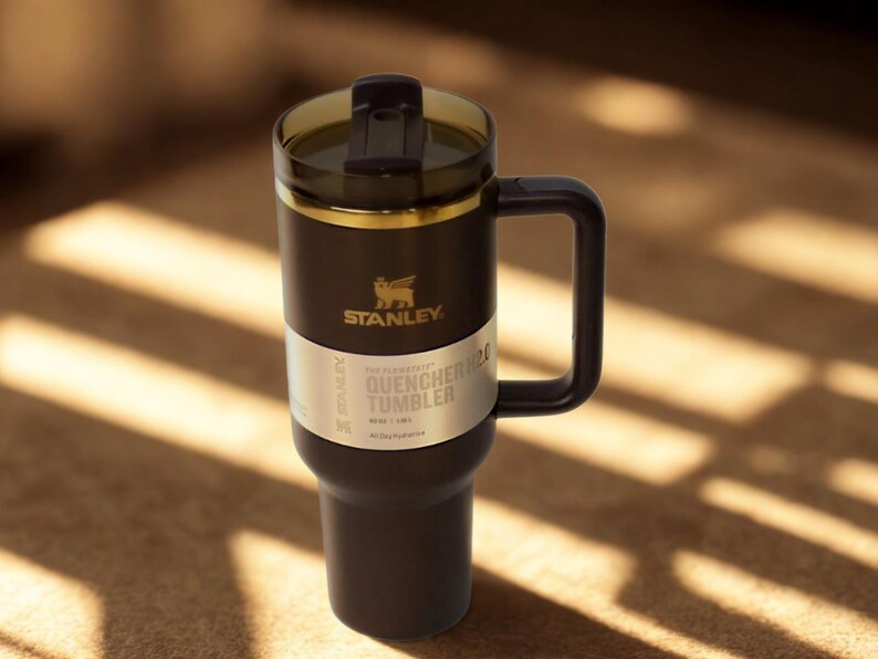 Może przedstawiać: Czarny kubek Stanley Quencher H2.0 z uchwytem i przezroczystą pokrywką. Kubek ma złoty pasek i logo Stanley. Tekst "QUENCHER TUMBLER" jest wydrukowany na białej etykiecie.