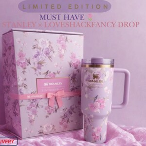 May include: A lavender Stanley tumbler with a floral pattern and a matching box featuring the text LIMITED EDITION MUST HAVE STANLEY x LOVESHACKFANCY DROP. The tumbler has a handle, a lid, and the Stanley logo printed on the front surface.