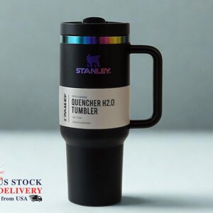 May include: Black Stanley Quencher H2.0 tumbler with a handle. The tumbler has a black lid with a rainbow-colored band and the Stanley logo. The text "QUENCHER H2.0 TUMBLER" is printed on a white label. The tumbler is designed to keep drinks cold.