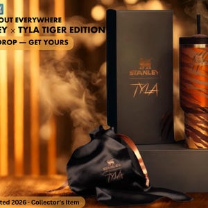 May include: A Stanley x Tyla Tiger Edition tumbler with a tiger stripe design, a black box, and a black drawstring bag. The tumbler has a handle and a straw. The text on the box reads "STANLEY" and "TYLA". The bag also has the "STANLEY" and "TYLA" logo.