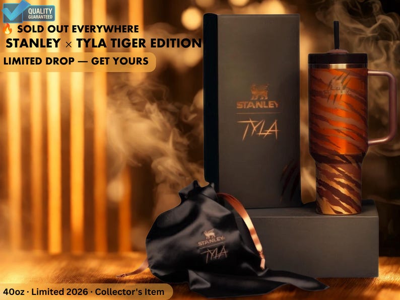 May include: A Stanley x Tyla Tiger Edition tumbler with a tiger stripe design, a black box, and a black drawstring bag. The tumbler has a handle and a straw. The text on the box reads "STANLEY" and "TYLA". The bag also has the "STANLEY" and "TYLA" logo.
