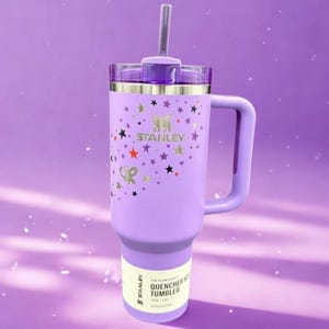 Op de afbeelding: Een lavendelkleurige Stanley Quencher-beker met een handvat en een rietje. De beker is versierd met gouden sterren en het Stanley-logo. De tekst "OR" is ook zichtbaar op de beker.