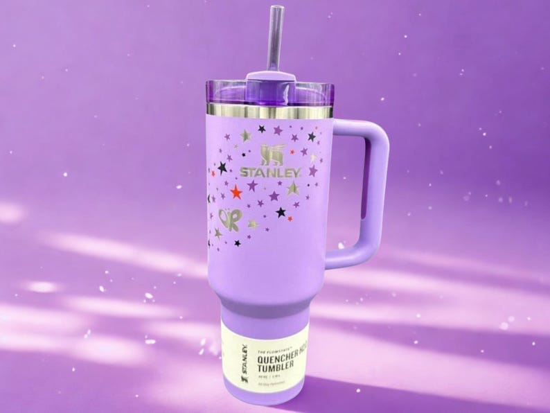 Op de afbeelding: Een lavendelkleurige Stanley Quencher-beker met een handvat en een rietje. De beker is versierd met gouden sterren en het Stanley-logo. De tekst "OR" is ook zichtbaar op de beker.