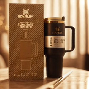Może przedstawiać: Czarny kubek Stanley Flowstate ze złotą pokrywką i uchwytem, obok brązowego pudełka. Pudełko zawiera rysunek kubka i logo Stanley. Kubek jest zaprojektowany tak, aby utrzymywać zimne napoje przez 11 godzin.
