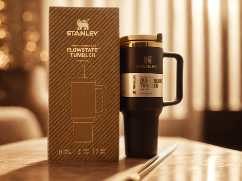 Może przedstawiać: Czarny kubek Stanley Flowstate ze złotą pokrywką i uchwytem, obok brązowego pudełka. Pudełko zawiera rysunek kubka i logo Stanley. Kubek jest zaprojektowany tak, aby utrzymywać zimne napoje przez 11 godzin.