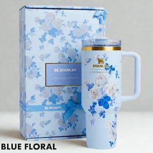 Puede incluir: Un vaso Stanley floral azul claro con asa y caja de regalo a juego. El vaso y la caja presentan un estampado floral azul y blanco. El vaso tiene un borde dorado y una pajita. El texto "STANLEY LOVESHACKFANCY" es visible.