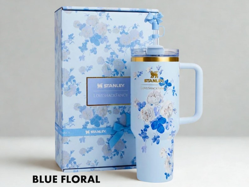 Puede incluir: Un vaso Stanley floral azul claro con asa y caja de regalo a juego. El vaso y la caja presentan un estampado floral azul y blanco. El vaso tiene un borde dorado y una pajita. El texto "STANLEY LOVESHACKFANCY" es visible.