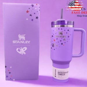 Op de afbeelding: Een lavendelkleurige Stanley Quencher H2.0 FlowState-beker met een handvat, rietje en deksel. De beker is versierd met gouden sterren en een Stanley-logo. Een bijpassende lavendelkleurige doos met het Stanley-logo en sterrenpatroon staat naast de beker.