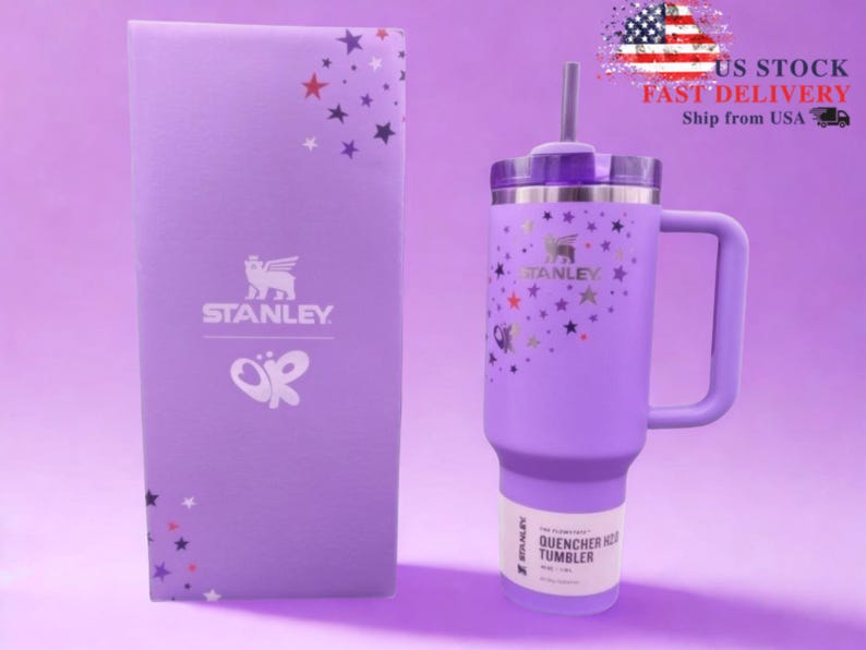 Op de afbeelding: Een lavendelkleurige Stanley Quencher H2.0 FlowState-beker met een handvat, rietje en deksel. De beker is versierd met gouden sterren en een Stanley-logo. Een bijpassende lavendelkleurige doos met het Stanley-logo en sterrenpatroon staat naast de beker.