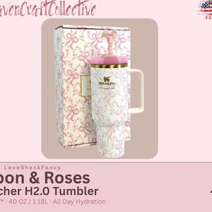 May include: A white Stanley Quencher H2.0 tumbler with a pink ribbon and rose pattern, featuring a matching pink lid, a handle, and a straw topper. The tumbler is displayed next to its original box, which shares the same pink and white ribbon design.