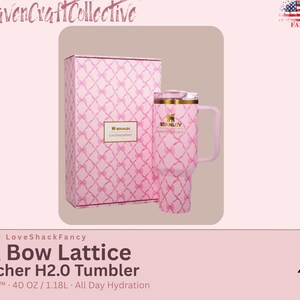 May include: A 40 oz Stanley x LoveShackFancy Quencher H2.0 tumbler and its matching box, both featuring a pink bow lattice pattern. The tumbler has a gold-tone rim, a handle, and a clear lid. The Stanley logo is printed on the front of the cup.