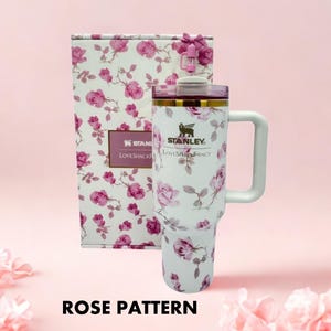 Puede incluir: Un vaso Stanley blanco con un estampado de rosas, un asa y una caja de regalo a juego. El vaso tiene una tapa morada y ribetes dorados. La caja de regalo tiene un lazo rosa y un estampado de rosas.