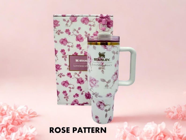 Puede incluir: Un vaso Stanley blanco con un estampado de rosas, un asa y una caja de regalo a juego. El vaso tiene una tapa morada y ribetes dorados. La caja de regalo tiene un lazo rosa y un estampado de rosas.