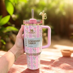 Może przedstawiać: Kubek Stanley Quencher H2.0 w kwiatowy wz&oacute;r z r&oacute;żowym uchwytem i pokrywką. Kubek ma słomkę i złoty kranowy dodatek. Widoczny jest tekst "LOVESHACKFANCY" i "QUENCHER TUMBLER".