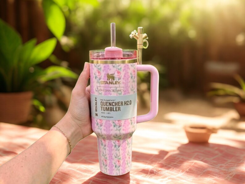 Może przedstawiać: Kubek Stanley Quencher H2.0 w kwiatowy wz&oacute;r z r&oacute;żowym uchwytem i pokrywką. Kubek ma słomkę i złoty kranowy dodatek. Widoczny jest tekst "LOVESHACKFANCY" i "QUENCHER TUMBLER".