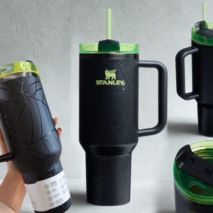 May include: A matte black Stanley tumbler featuring a subtle spiderweb pattern, a sturdy side handle, and a translucent neon green lid with a matching straw. The cup displays the Stanley logo in neon green and includes a black sliding lid mechanism.