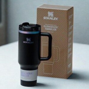 May include: A black Stanley Flowstate tumbler with a handle and a matching box. The tumbler has a black lid with a silver rim and the Stanley logo. The box is brown with the Stanley logo and product information.