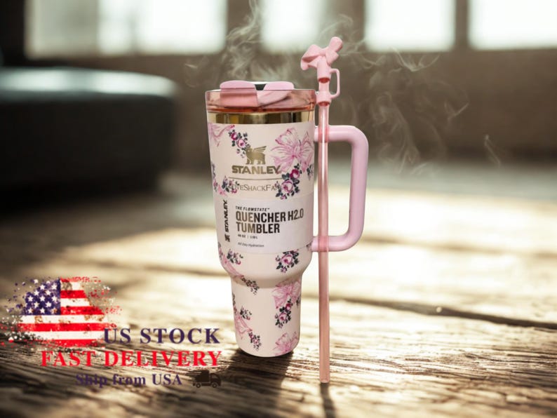 Puede incluir: Un vaso Stanley Quencher H2.0 blanco con un dise&ntilde;o floral en rosa y dorado. El vaso tiene un asa, pajita y tapa rosas. Se puede leer el texto "STANLEY" y "QUENCHER H2.0 TUMBLER".