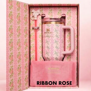 Puede incluir: Un termo Stanley rosa con un estampado floral, una pajita a juego y una bolsa de terciopelo rosa, presentado en una caja decorativa. La caja y el termo muestran el texto "Ribbon Rose".