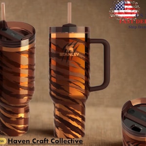 May include: Two Stanley brand travel mugs with a tiger stripe pattern in shades of brown and orange. Each mug has a handle, a lid, and a straw. The mugs are displayed on a brown background. The text "Haven Craft Collective" is at the bottom.