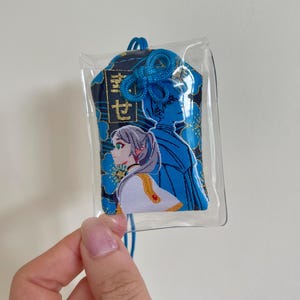 May include: A blue and black embroidered charm with a clear plastic covering. The charm features two anime-style figures and Japanese text. The design includes blue floral accents and a blue cord for hanging.
