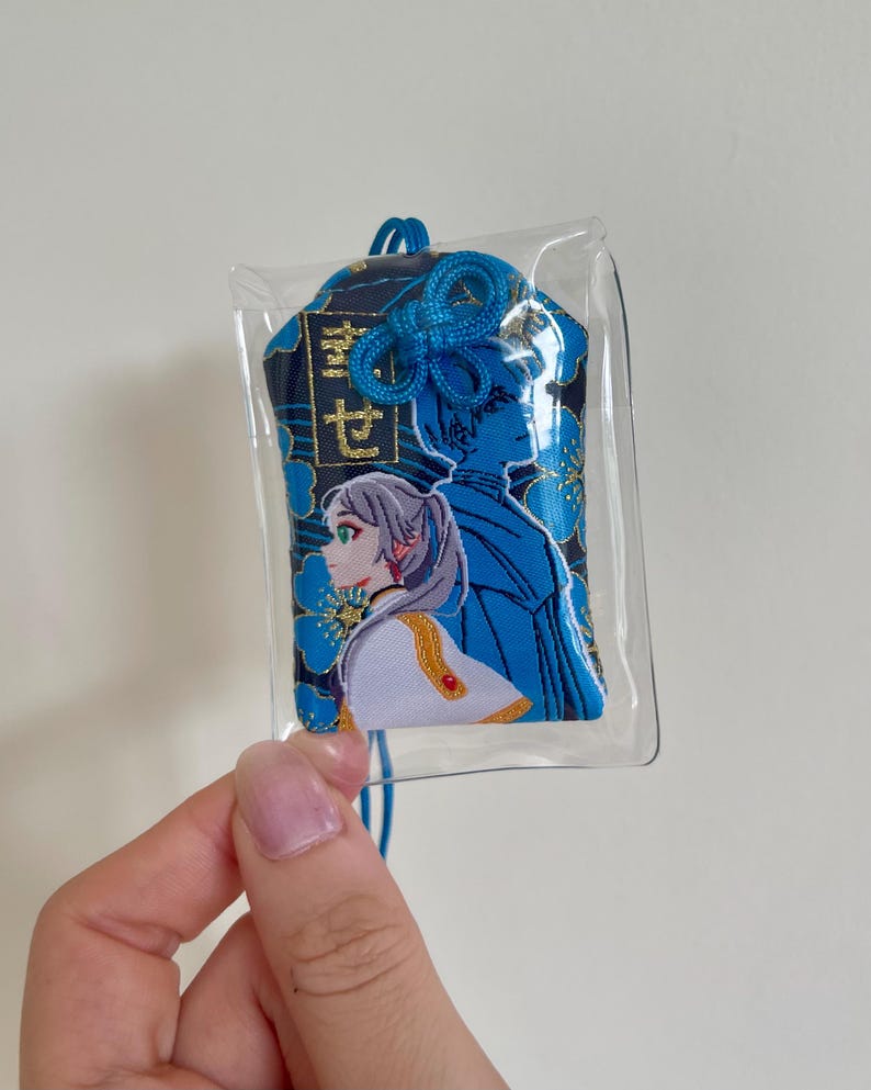 May include: A blue and black embroidered charm with a clear plastic covering. The charm features two anime-style figures and Japanese text. The design includes blue floral accents and a blue cord for hanging.