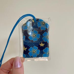 May include: A small, navy blue pouch with a floral pattern in shades of blue and gold, encased in a clear plastic sleeve. A blue cord is attached for carrying. The pouch features a repeating design of stylized flowers and star shapes.