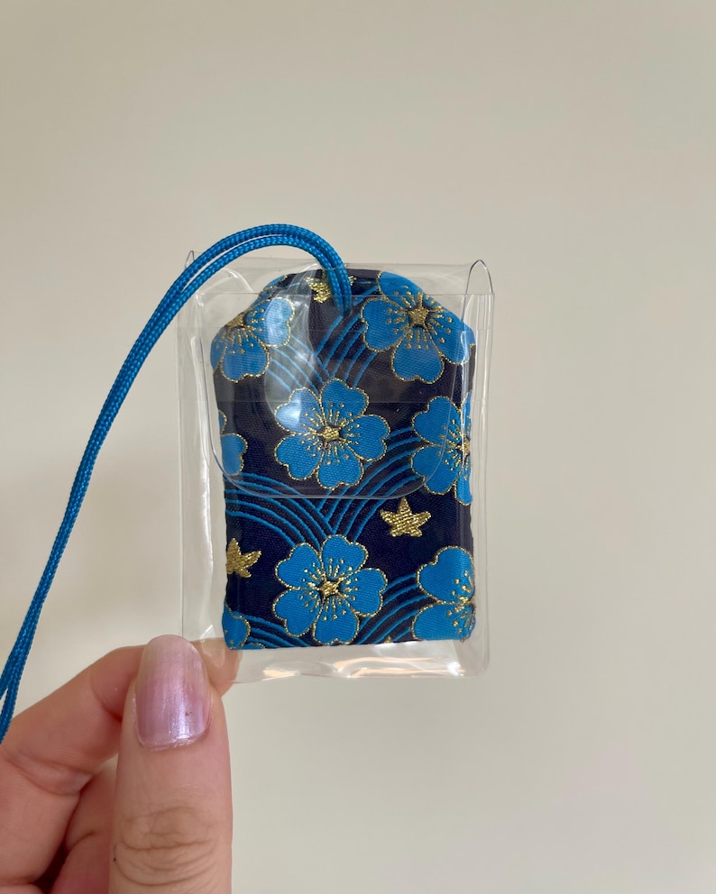 May include: A small, navy blue pouch with a floral pattern in shades of blue and gold, encased in a clear plastic sleeve. A blue cord is attached for carrying. The pouch features a repeating design of stylized flowers and star shapes.