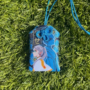 May include: A blue and black embroidered charm with a detailed illustration of a character with gray hair and a white and gold outfit. The charm has a blue cord and a decorative knot at the top. The background is a field of green grass.