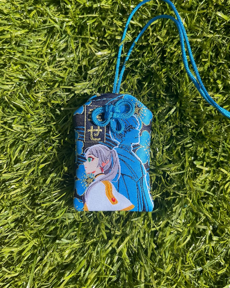 May include: A blue and black embroidered charm with a detailed illustration of a character with gray hair and a white and gold outfit. The charm has a blue cord and a decorative knot at the top. The background is a field of green grass.