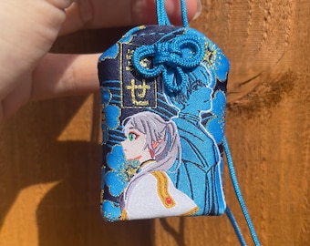 May include: Embroidered blue and navy charm with a detailed anime-style illustration of two figures. The charm features a blue cord and a decorative knot. The design includes floral patterns and Japanese text.