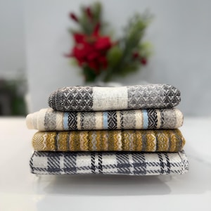 May include: A stack of four woven blankets in various patterns. The top blanket is gray and white with a diamond pattern. The second blanket is beige and gray with a striped pattern. The third blanket is yellow and brown with a striped pattern. The bottom blanket is gray and white with a plaid pattern.