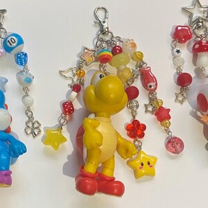 May include: Three keychains featuring cartoon characters. One keychain has a blue Yoshi figure, another has a yellow Koopa Troopa, and the third has a Toad figure. Each keychain has a star-shaped clasp and is adorned with beads and charms.
