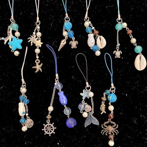 May include: Ten colorful phone charms with a nautical theme. The charms feature seashells, starfish, mermaids, anchors, and other ocean-inspired designs. The charms are strung on thin cords with white beads.