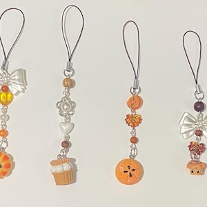 Op de afbeelding: Vier decoratieve telefoonbedels met verschillende ontwerpen. Eén heeft een witte strik, een oranje bloem en kralen. Een andere heeft een bloem, hartjes en een cupcake. De derde heeft oranje kralen en een vrucht. De laatste heeft een strik en een cupcake.