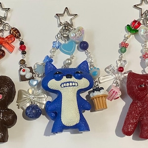 May include: Three quirky keyrings with star-shaped clasps. One is brown, one is blue, and one is red. Each features a unique character and decorative beads, bows, and charms. The keyrings have a fun, playful design.
