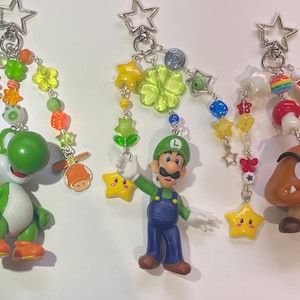 May include: Three keychains featuring characters from the Super Mario video game series. Each keychain has a star-shaped clasp and a beaded chain with a character figurine. The characters include Yoshi, Luigi, and a Goomba.