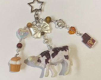 May include: A whimsical keychain featuring a white and brown cow figurine. The keychain includes a star-shaped clasp, a white bow, a heart, a cupcake, and a chocolate bar charm, all connected with beads. The cow figurine is the focal point.