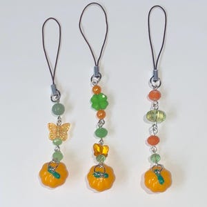 May include: Three decorative charms with orange pumpkin pendants, butterfly and clover beads, and green and orange accents. Each charm has a silver-coloured metal loop and a black cord for attaching to a phone or bag.