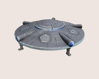 May include: A silver and blue flying saucer with a round, flat bottom and three legs. The saucer has a circular window in the center and four smaller windows around the edge.