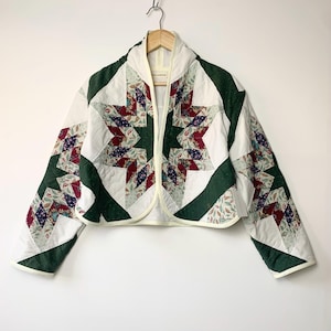 May include: A quilted jacket with a starburst pattern. The jacket has a white background with green and patterned fabric accents in red, blue, and green. It is hanging on a wooden hanger.