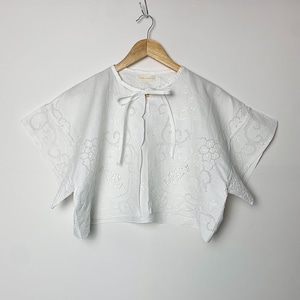 May include: White cotton blouse with a scalloped neckline and embroidered floral details. The blouse has a tie at the neck and short sleeves.