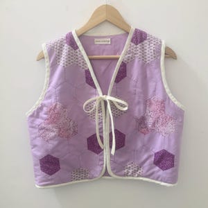 Pode incluir: Um colete acolchoado em tons de lavanda e roxo, com um padrão geométrico de hexágonos e detalhes florais. O colete tem uma borda branca e um fecho de laço. A etiqueta diz "maze workshop".
