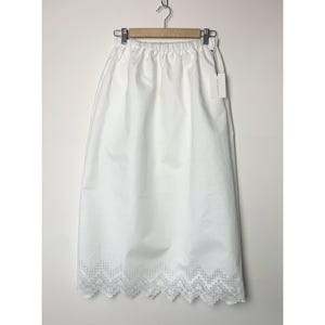 May include: A white skirt with an elastic waistband and a delicate lace trim along the hemline. The skirt is hanging on a wooden hanger against a white background. A tag is attached to the skirt.